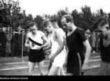 Archiwalne zdjęcie biegaczy na stadionie przed startem, w tle kibice przy ogrodzeniu.