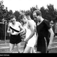 Archiwalne zdjęcie biegaczy na stadionie przed startem, w tle kibice przy ogrodzeniu.