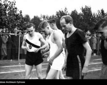 Archiwalne zdjęcie biegaczy na stadionie przed startem, w tle kibice przy ogrodzeniu.