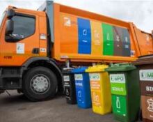 Nowoczesna, pomarańczowa śmieciarka MPO Warszawa z kolorowymi panelami segregacji. Przed pojazdem stoi zestaw pięciu kolorowych pojemników na odpady: czarny, niebieski, żółty, zielony i brązowy BIO. Wszystkie kontenery mają czytelne etykiety MPO i ikony segregacji.