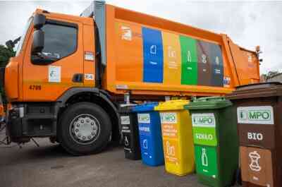Nowoczesna, pomarańczowa śmieciarka MPO Warszawa z kolorowymi panelami segregacji. Przed pojazdem stoi zestaw pięciu kolorowych pojemników na odpady: czarny, niebieski, żółty, zielony i brązowy BIO. Wszystkie kontenery mają czytelne etykiety MPO i ikony segregacji.