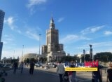 Pałac Kultury i Nauki w Warszawie widziany od strony Placu Defilad — widok od poziomu ulicy, na pierwszym planie przechodnie i parking, w tle charakterystyczna wieża PKiN na tle błękitnego nieba.