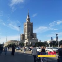 Pałac Kultury i Nauki w Warszawie widziany od strony Placu Defilad — widok od poziomu ulicy, na pierwszym planie przechodnie i parking, w tle charakterystyczna wieża PKiN na tle błękitnego nieba.