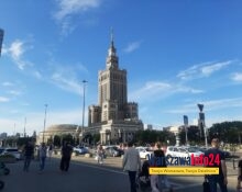 Pałac Kultury i Nauki w Warszawie widziany od strony Placu Defilad — widok od poziomu ulicy, na pierwszym planie przechodnie i parking, w tle charakterystyczna wieża PKiN na tle błękitnego nieba.