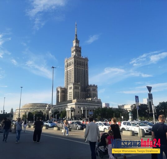 Pałac Kultury i Nauki w Warszawie widziany od strony Placu Defilad — widok od poziomu ulicy, na pierwszym planie przechodnie i parking, w tle charakterystyczna wieża PKiN na tle błękitnego nieba.