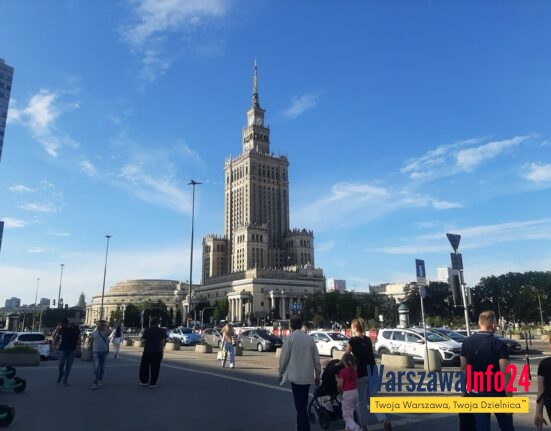 Pałac Kultury i Nauki w Warszawie widziany od strony Placu Defilad — widok od poziomu ulicy, na pierwszym planie przechodnie i parking, w tle charakterystyczna wieża PKiN na tle błękitnego nieba.