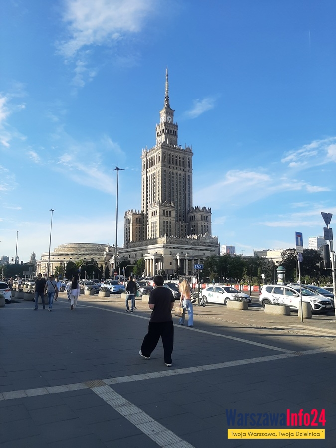 Pałac Kultury i Nauki w Warszawie — widok od strony Alej Jerozolimskich. Na pierwszym planie przechodnie, rodziny z wózkami i ruch uliczny, w tle sylwetka PKiN na tle błękitnego nieba.